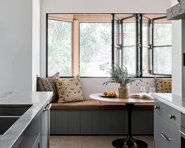 Cozy kitchen nook with built-in banquette seating, patterned cushions, a round table, flowers, and open windows. Blue joinery in a galley shaped kitchen is just visible in the foreground.