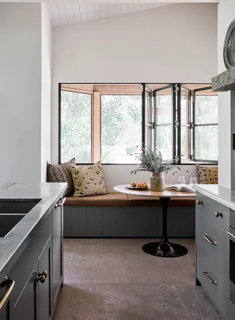 Cozy kitchen nook with built-in banquette seating, patterned cushions, a round table, flowers, and open windows. Blue joinery in a galley shaped kitchen is just visible in the foreground.