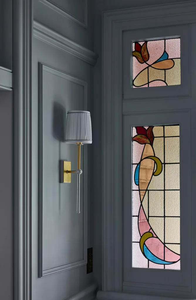 Wall sconce with pleated shade beside colorful stained glass window in a room with dark paneled walls.