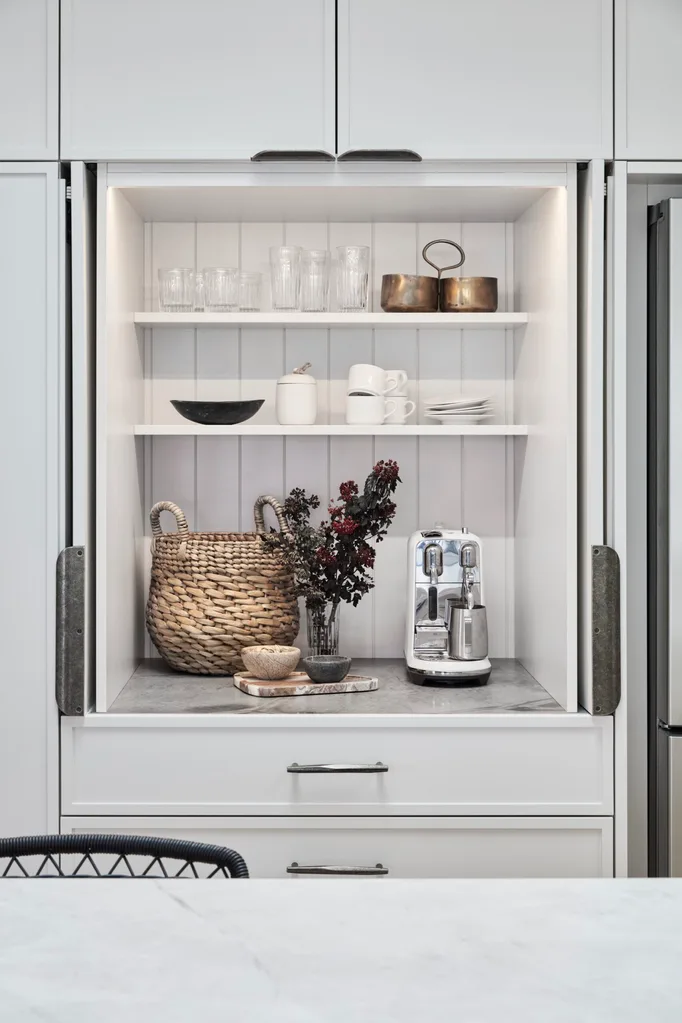 Modern kitchen coffee station with glasses, copper pots, wicker basket, coffee maker, and flowers on marble benchtop.