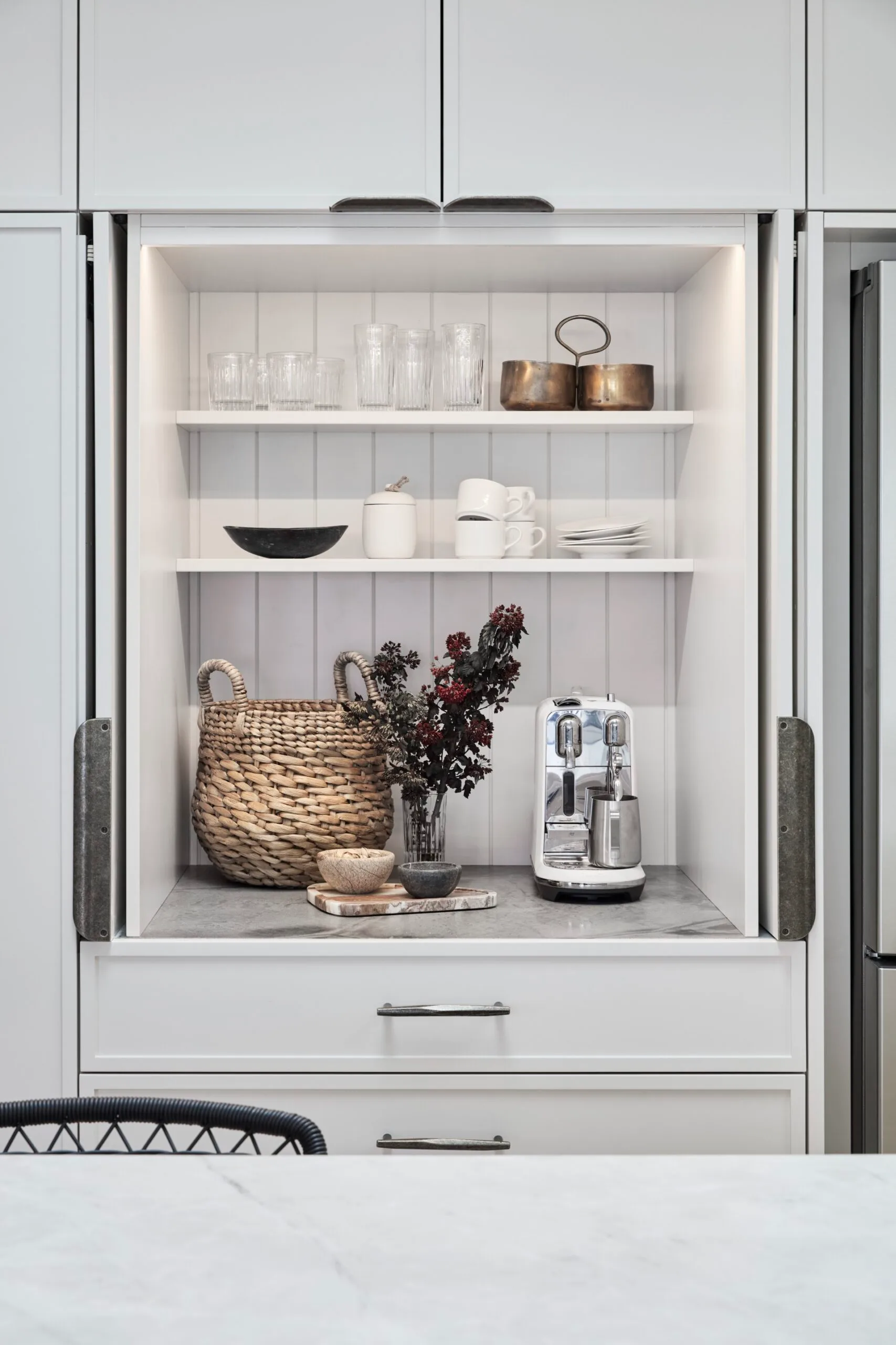 Modern kitchen coffee station with glasses, copper pots, wicker basket, coffee maker, and flowers on marble benchtop.