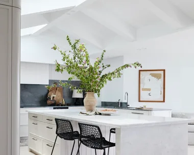 Bright kitchen with white island, vase of green branches, two black chairs, and a framed wall art.
