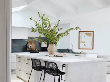 Bright kitchen with white island, vase of green branches, two black chairs, and a framed wall art.
