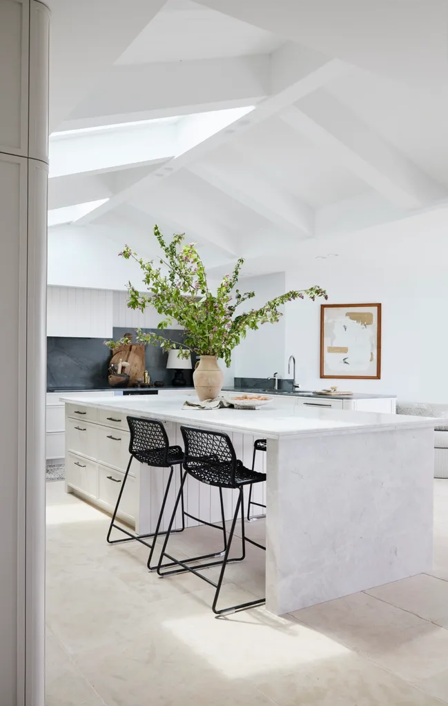 Bright kitchen with white island, vase of green branches, two black chairs, and a framed wall art.