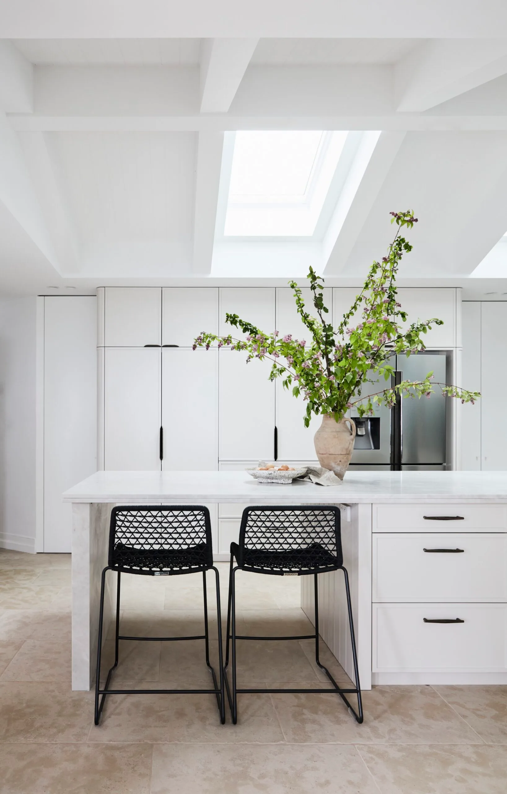 Bright modern kitchen with white cabinetry, a large island, two black stools, and a vase of green branches under a skylight in a pitched ceiling.