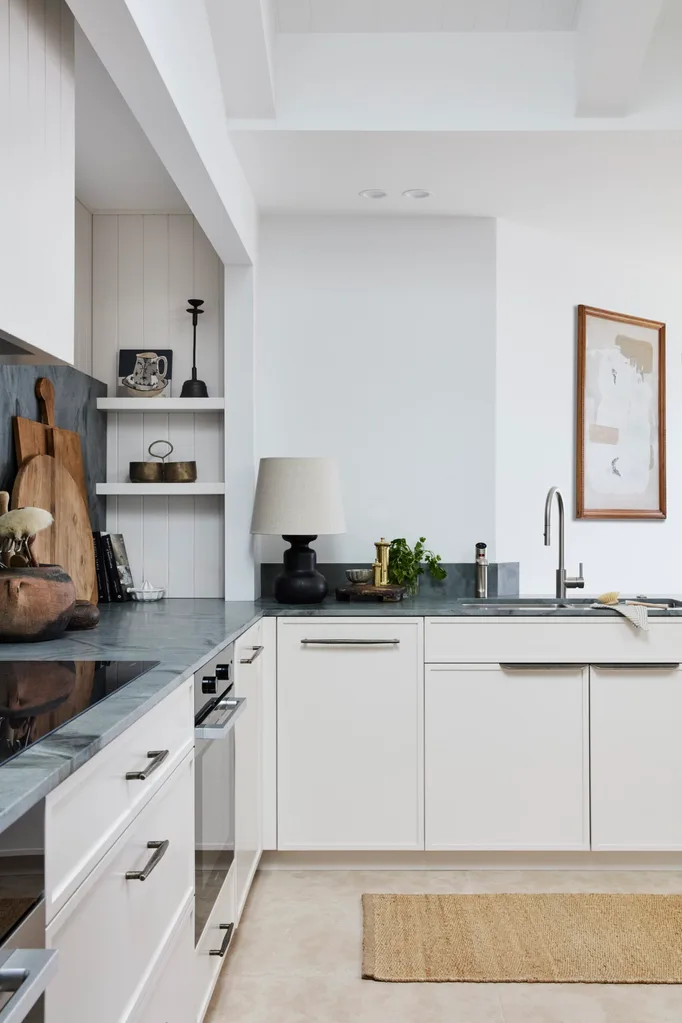 Modern kitchen with white cabinets, grey stone benchtops. black lamp, timber chopping boards, herbs, and framed abstract art on the wall.