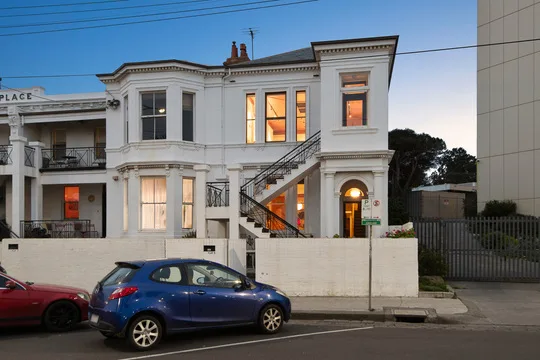 Victorian-style white building with lit windows and external staircase; blue and red cars parked in front Ron Barassi St Kilda home