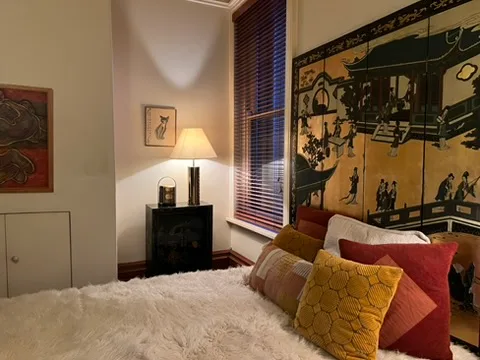 Cozy bedroom with a large Asian-themed screen, plush pillows on a bed, and a soft lamp glow beside a window with blinds.