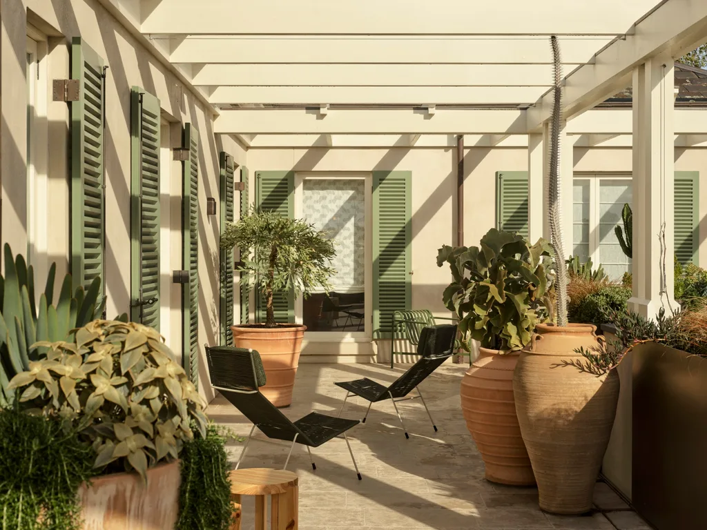 Outdoor patio with green shutters, large terracotta pots with plants, and black chairs under a white pergola.