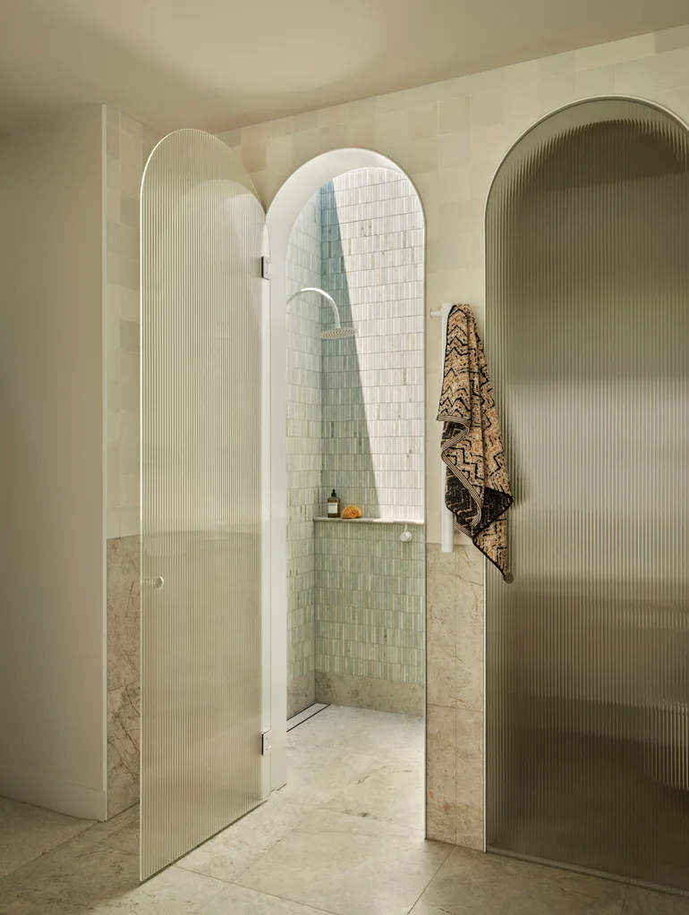 Archway shower with glass door, beige tiles, and a hanging towel. Bottle and sponge on shower shelf.