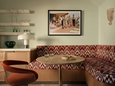 Retro dining nook with patterned banquette, round table, orange chair, and wall art depicting classic beach scene.