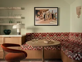 Retro dining nook with patterned banquette, round table, orange chair, and wall art depicting classic beach scene.
