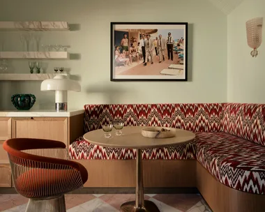Retro dining nook with patterned banquette, round table, orange chair, and wall art depicting classic beach scene.