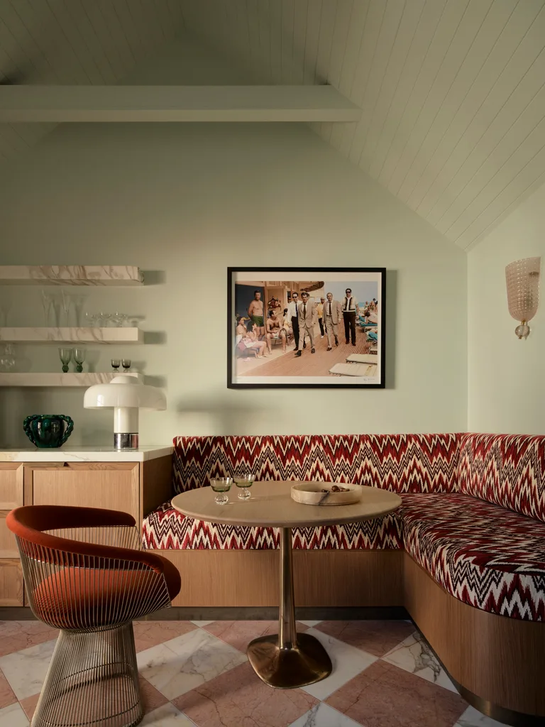 Retro dining nook with patterned banquette, round table, orange chair, and wall art depicting classic beach scene.