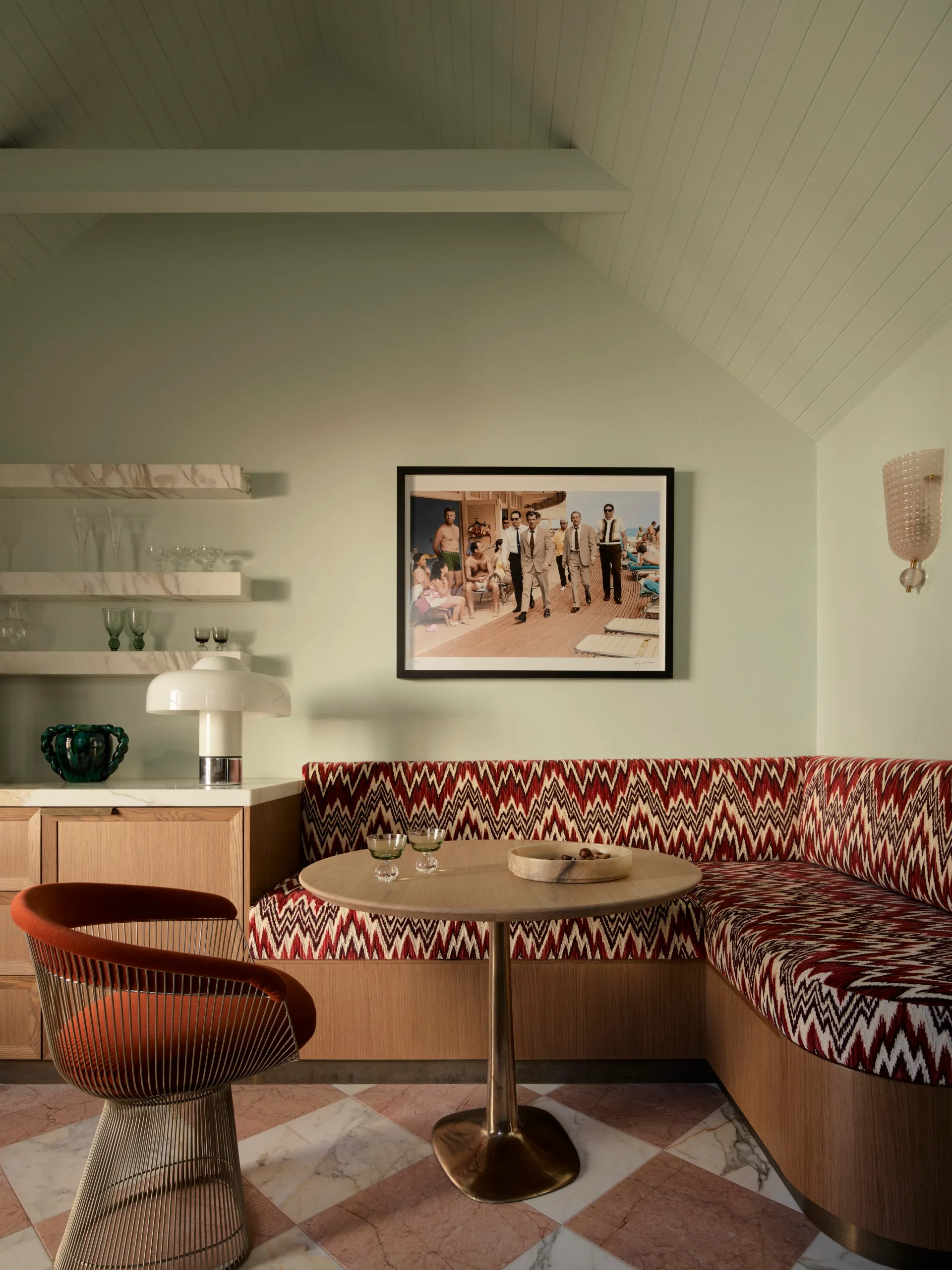 Retro dining nook with patterned banquette, round table, orange chair, and wall art depicting classic beach scene.