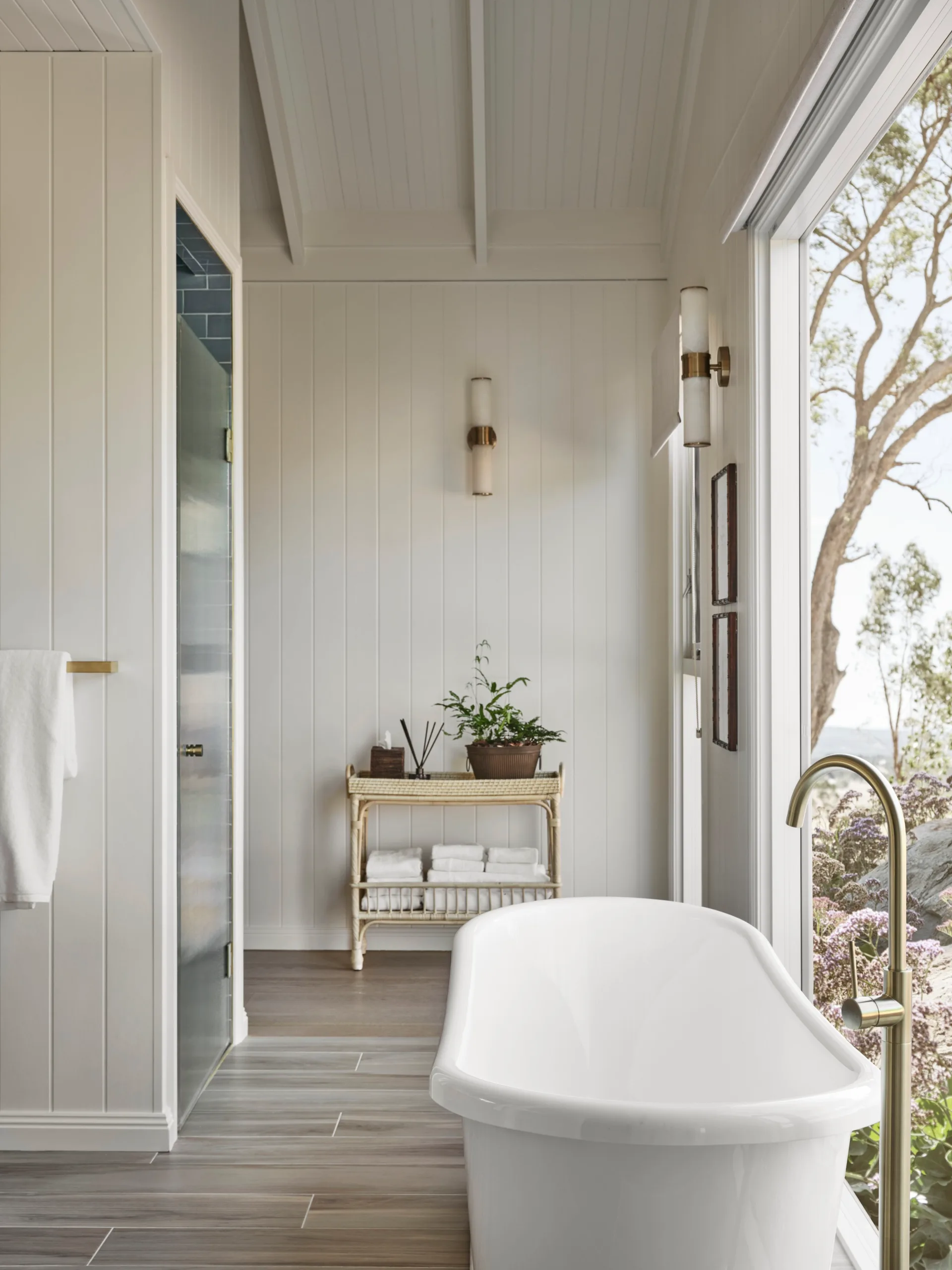 Bright bathroom with a white freestanding tub, wooden shelves, potted plant, and large window with garden view.