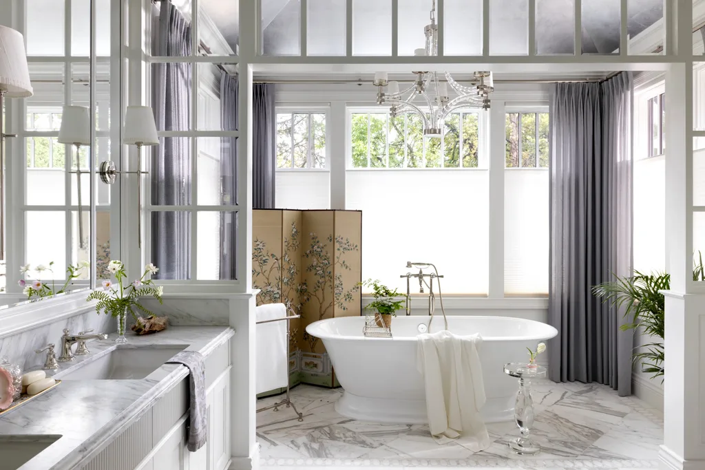 Elegant bathroom with white freestanding tub, marble surfaces, gray curtains, chandelier, and floral screen.