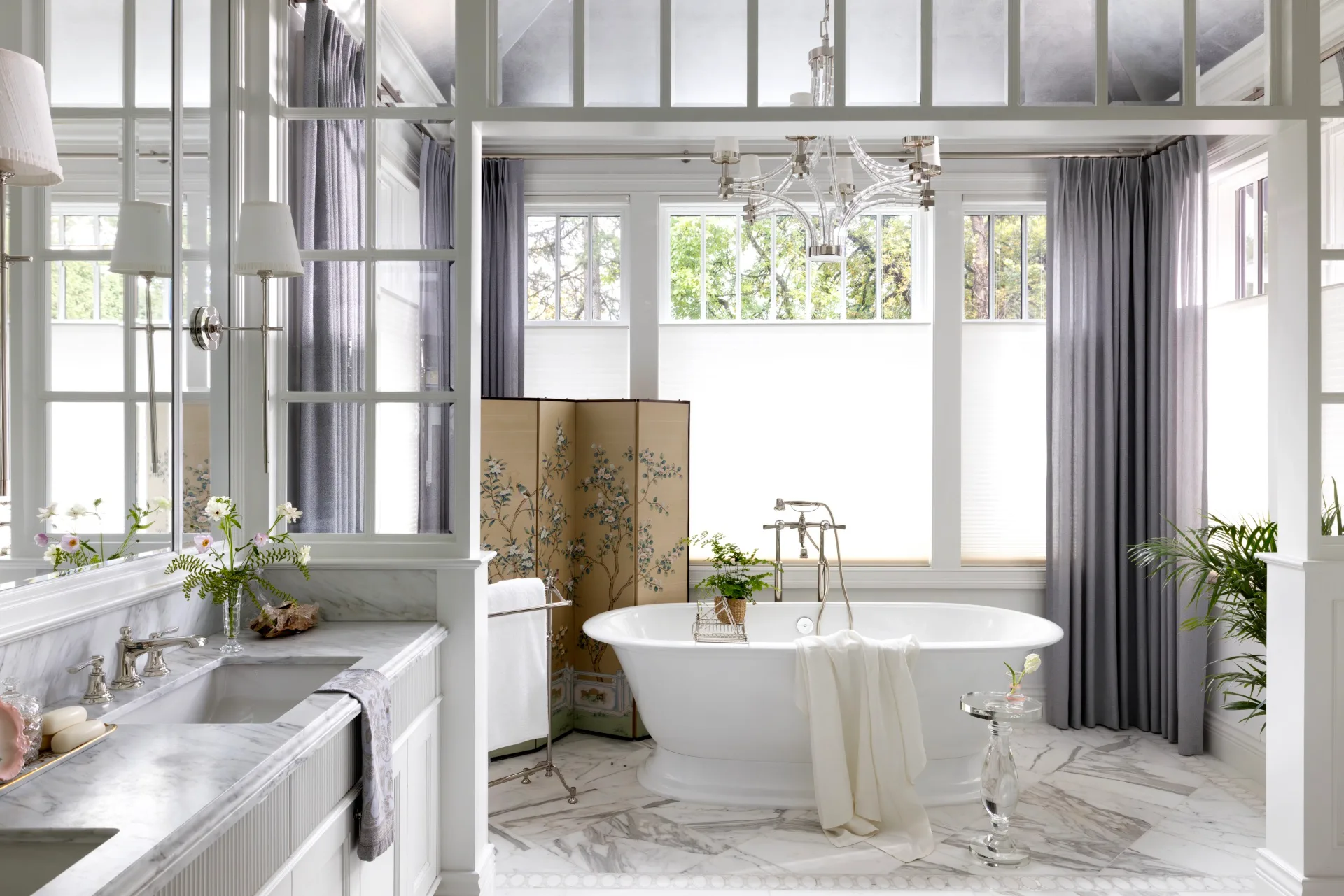 Elegant bathroom with white freestanding tub, marble surfaces, gray curtains, chandelier, and floral screen.