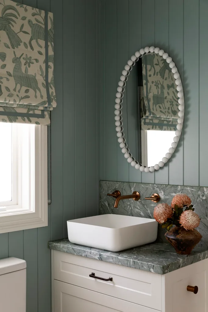Elegant bathroom with oval mirror, white sink, gold faucet, textured wall, floral vase, and patterned blinds.