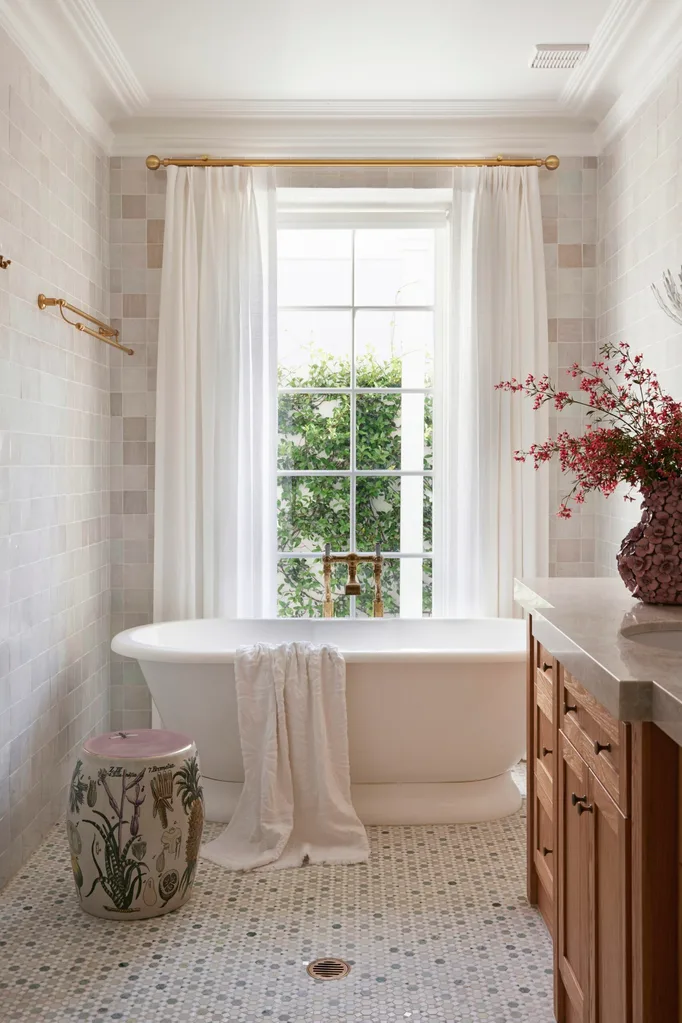 Elegant bathroom with white curtains, freestanding bathtub, floral stool, and pink flowers in vase on tiled floor.