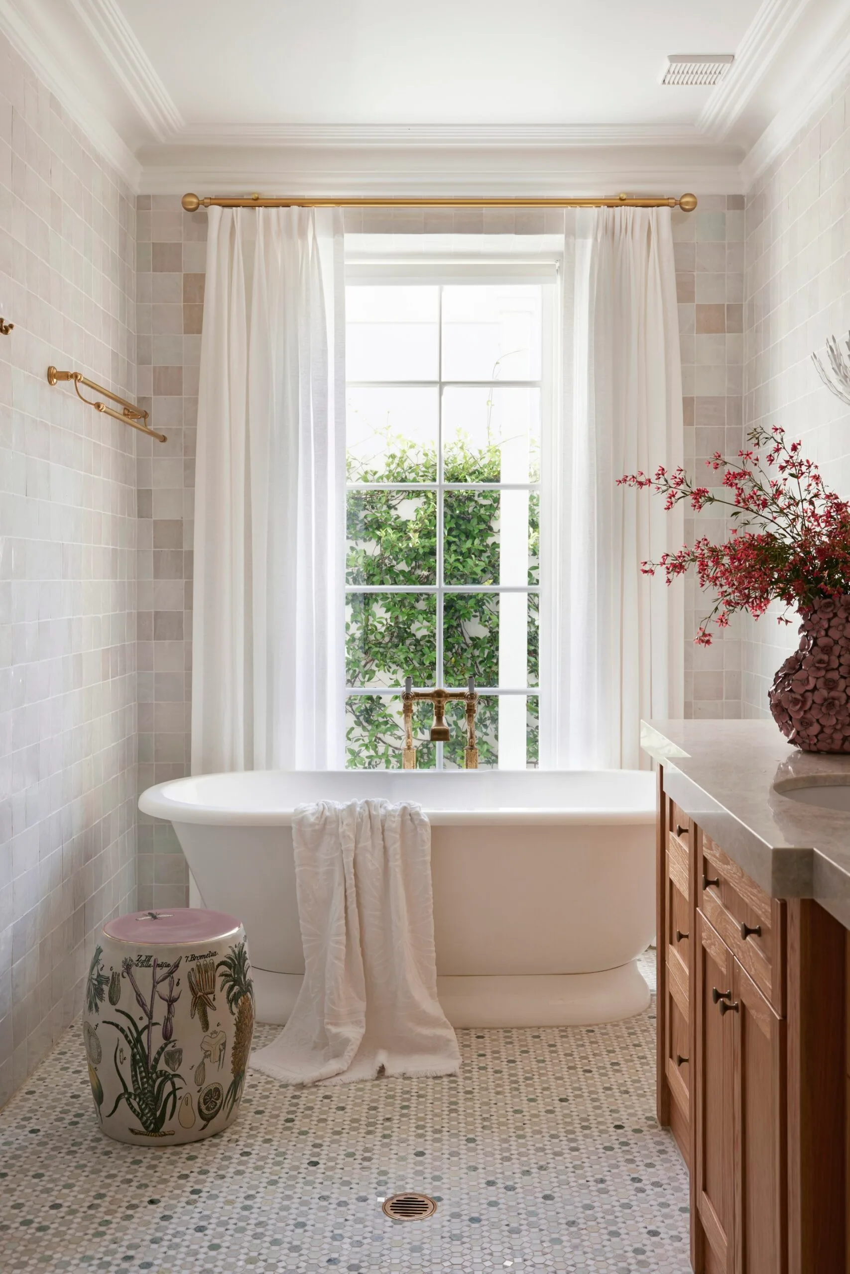 Elegant bathroom with white curtains, freestanding bathtub, floral stool, and pink flowers in vase on tiled floor.