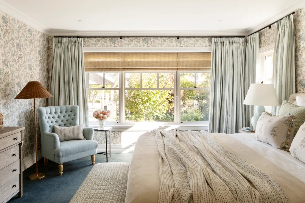 Cozy bedroom with floral wallpaper, light blue chair, large window with curtains, and bed with patterned pillows.