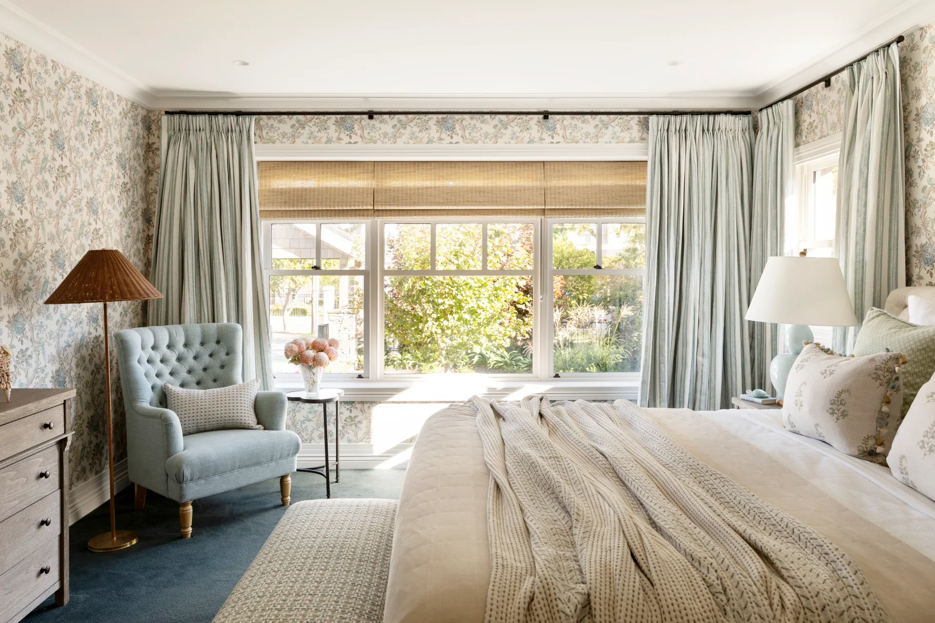 Cozy bedroom with floral wallpaper, light blue chair, large window with curtains, and bed with patterned pillows.