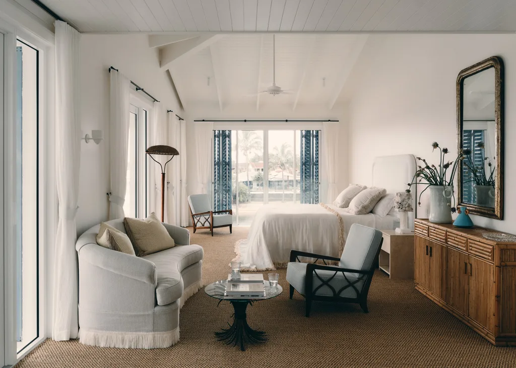 Cozy bedroom with neutral decor, white bed, cushioned chairs, a sofa, and large windows overlooking a patio.