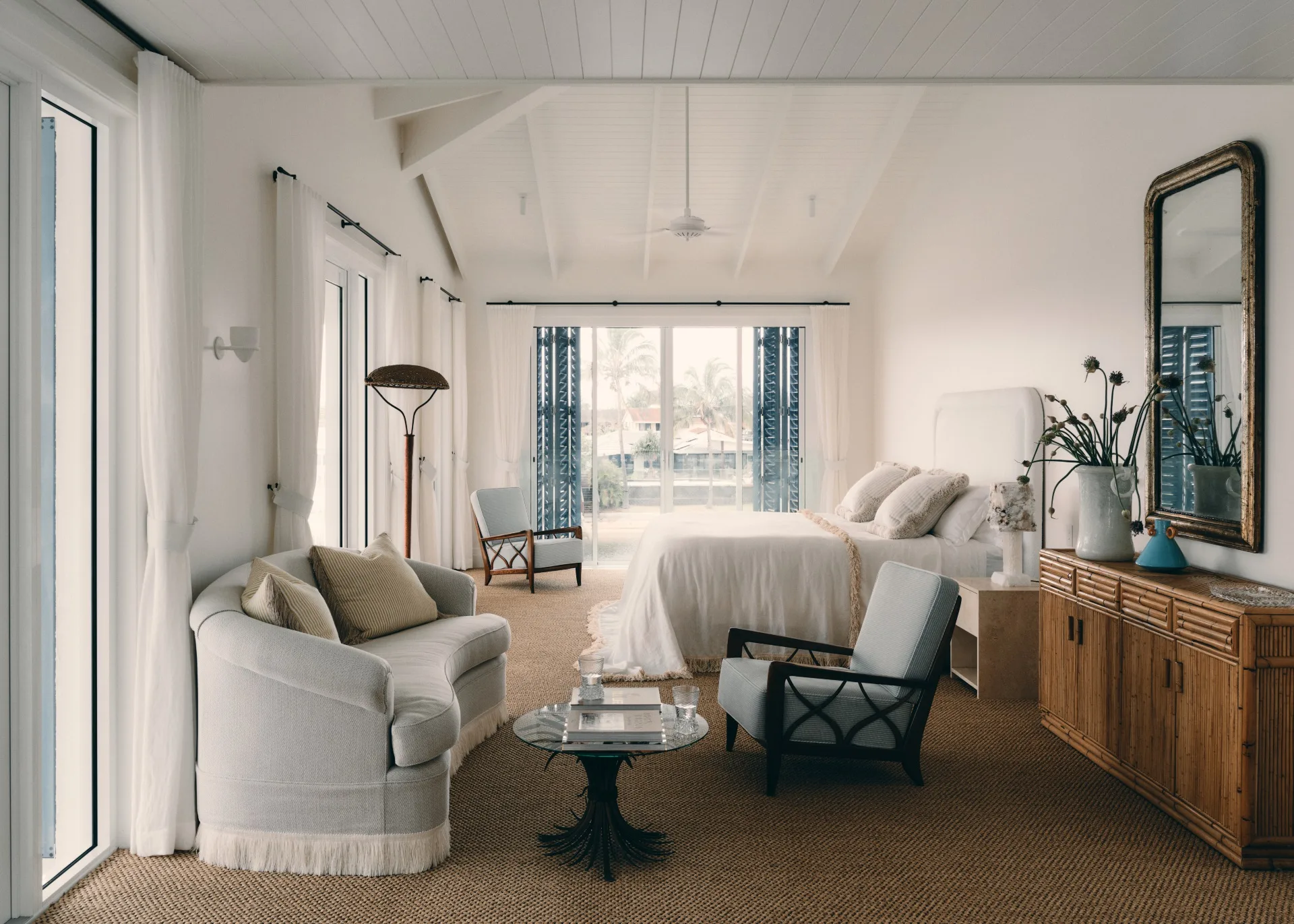 Cozy bedroom with neutral decor, white bed, cushioned chairs, a sofa, and large windows overlooking a patio.