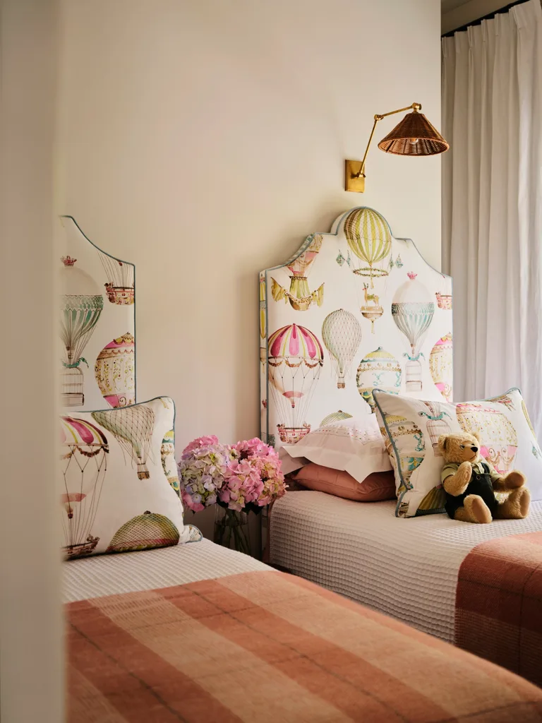 Two twin beds with balloon-themed headboards, pink plaid blankets, pillows, a teddy bear, and a vase of flowers.
