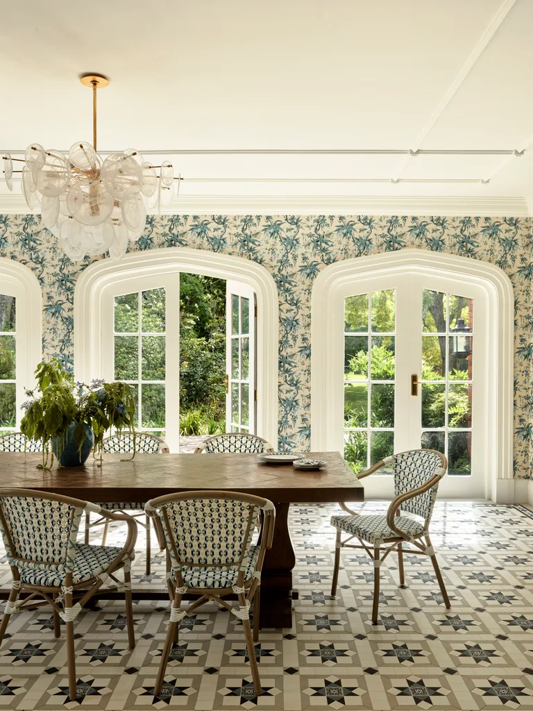 Elegant dining room with patterned floor, large wooden table, decorative chairs, floral wallpaper, and glass doors to garden.