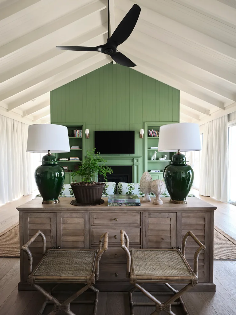 Cozy living room with green paneled wall, black ceiling fan, wooden furniture, large lamps, and a potted plant.