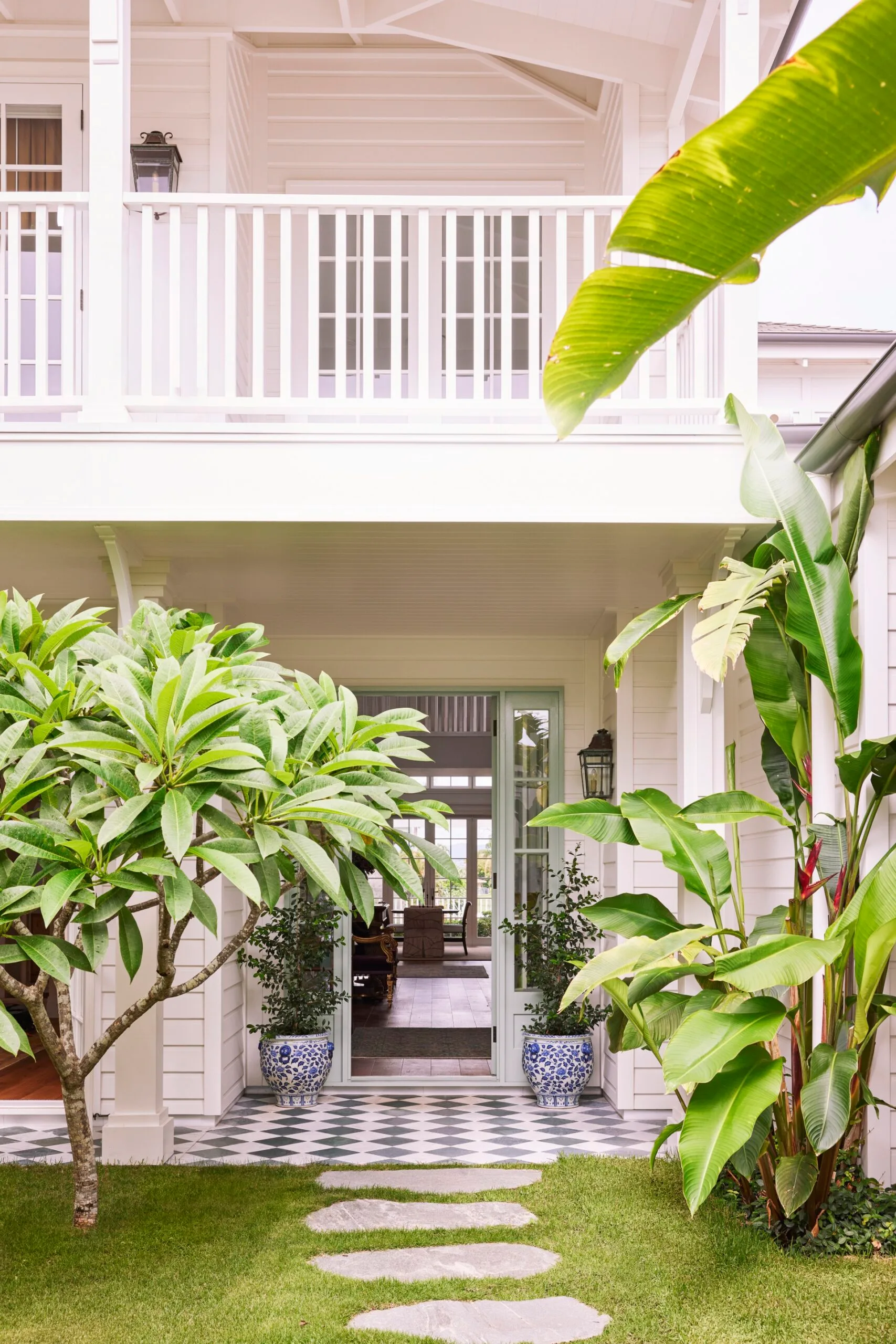 Elegant tropical patio with green plants, a pathway, and an open door leading to a stylish white-panelled home interior.
