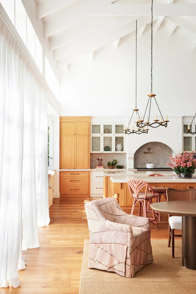 Bright kitchen with exposed beams, natural wood cabinets, island, dining set, chandelier, large windows, and curtains.