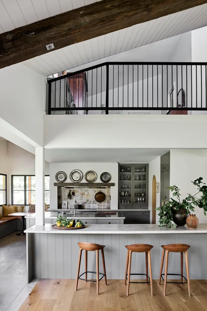 Modern kitchen with wooden stools at island, plants, stone backsplash, and upper loft with railing.