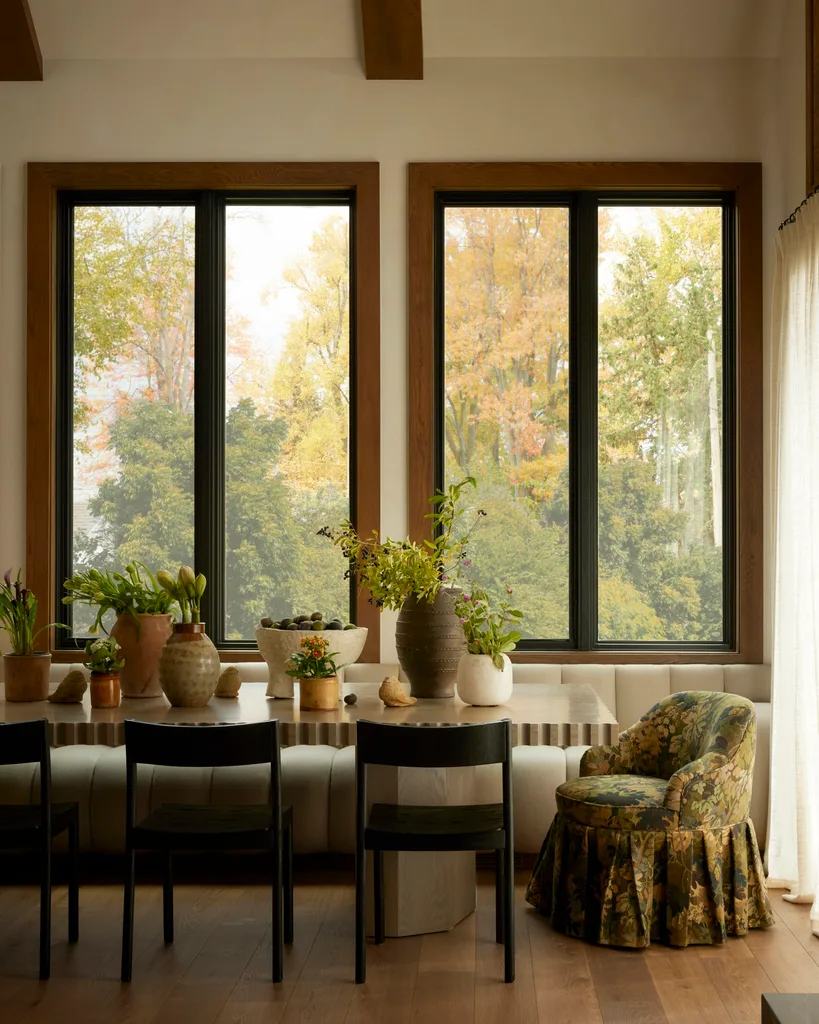 Cozy dining area with wooden table, plants in pots, black chairs, and a floral-patterned armchair by large windows.