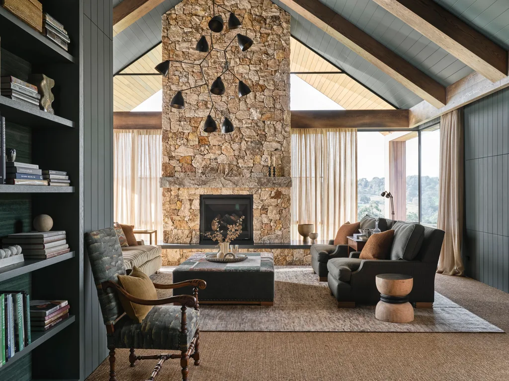 Luxurious living room with stone fireplace, dark wood beams, modern chandelier, and cozy seating area by large windows.