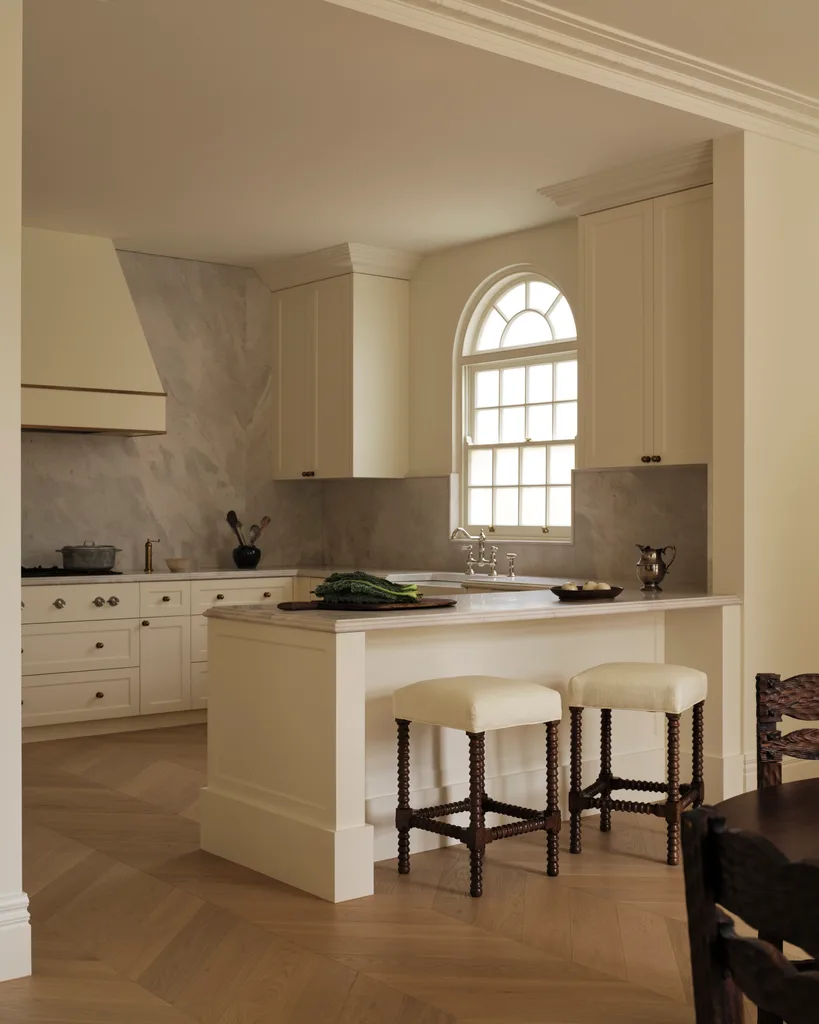 Warm, elegant kitchen with cream cabinetry, marble backsplash, arched window, and two cushioned stools at an island.