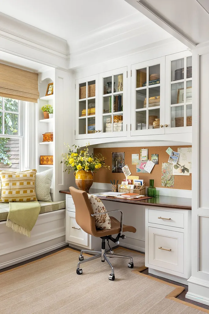 Cozy home office with white cabinets, a corkboard, yellow flowers, a window seat, and a swivel chair.