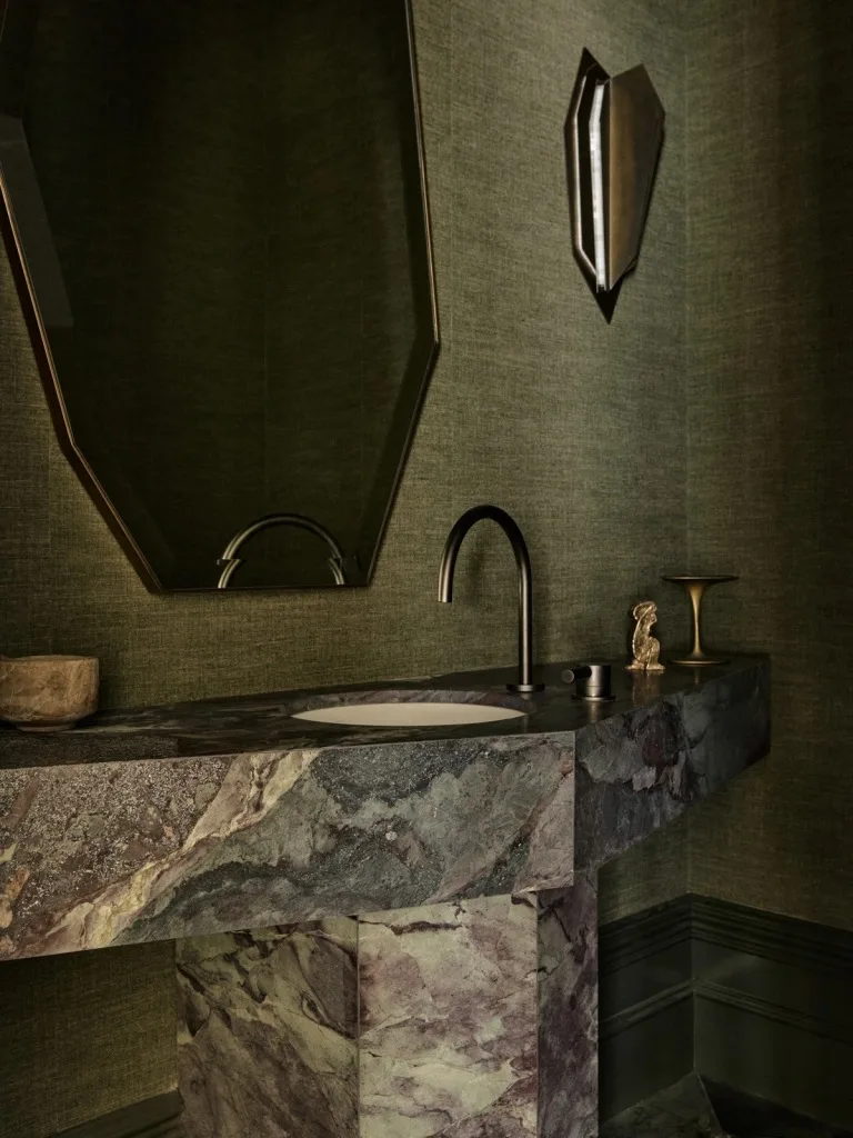 Marble sink in a modern bathroom with green textured walls, angular mirror, and elegant lighting fixture.
