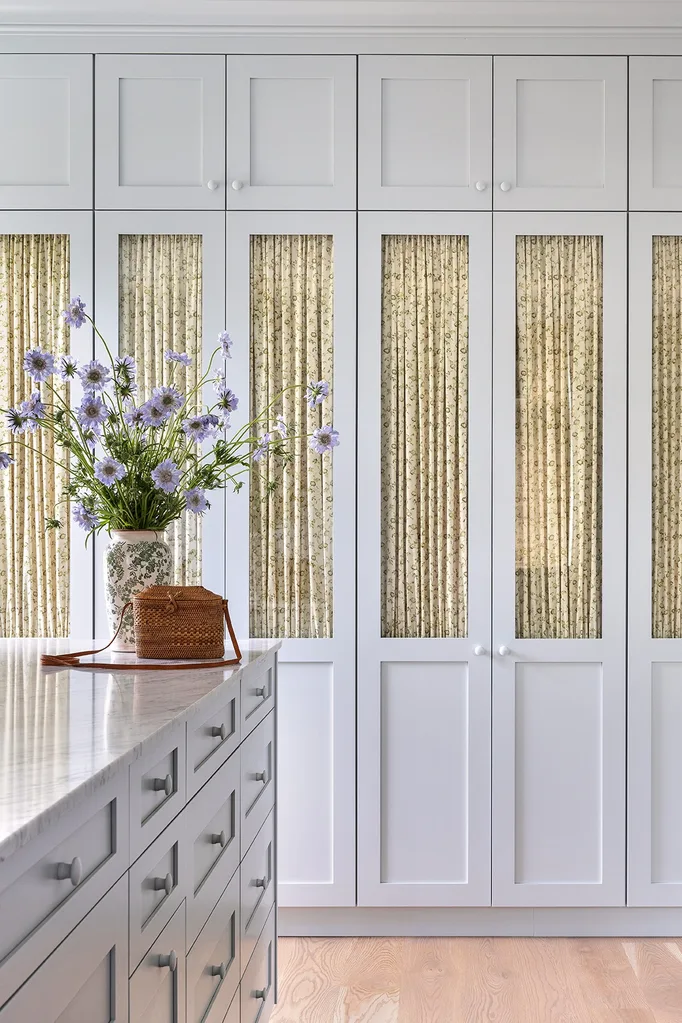 Kitchen with white cabinets, floral curtains, marble countertop, vase of purple flowers, and a woven basket on the counter.