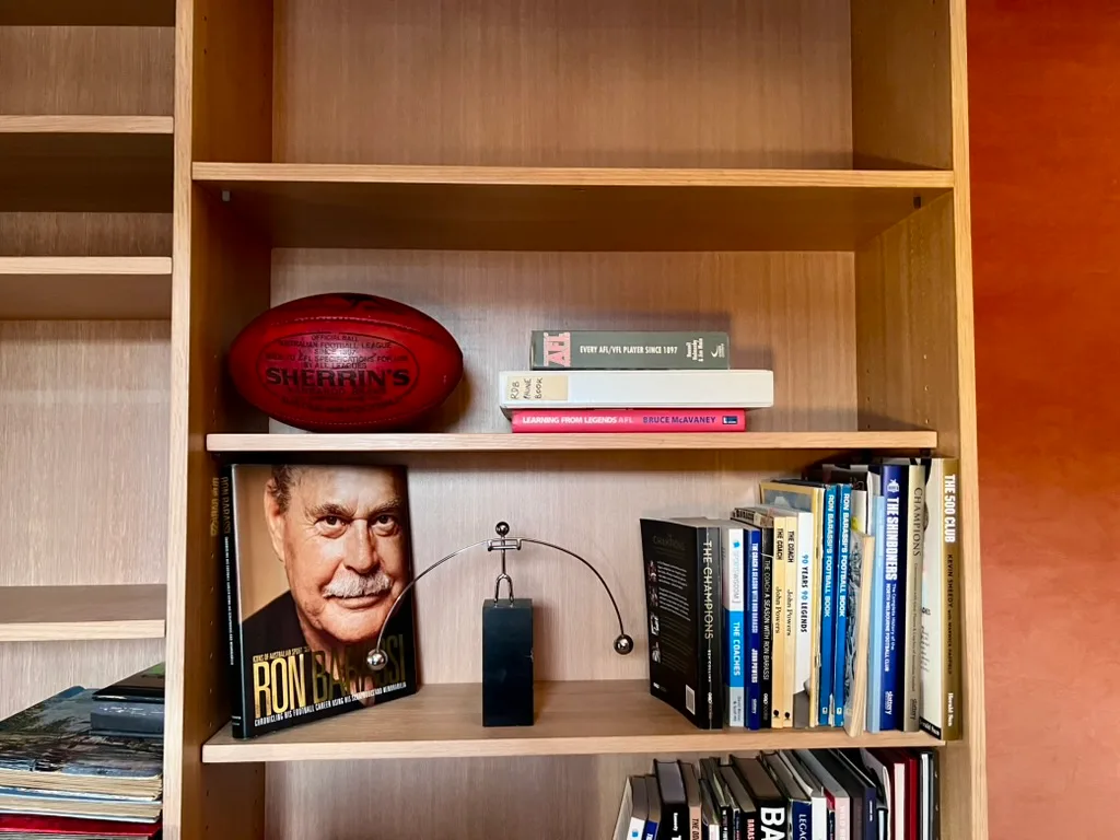 Wooden bookshelf with books, a red Sherrin football, and Ron Barassi's book displayed prominently Ron Barassi St Kilda home