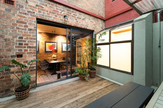 Outdoor patio with wooden flooring, potted plants, and view into cozy room with brick wall, art, and seating area through glass doors.