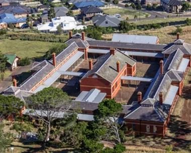 Aerial view of a historic bluestone building complex with orange rooftops, surrounded by trees and suburban houses. The Block 2027 location.