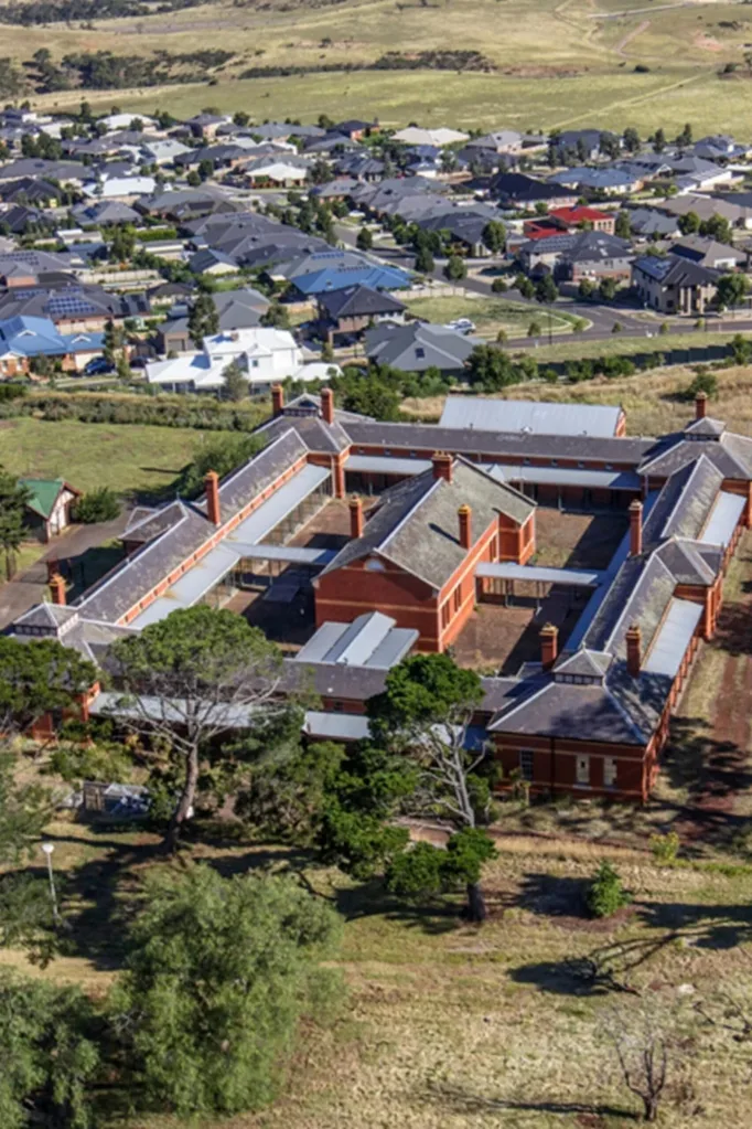 Aerial view of a historic bluestone building complex with orange rooftops, surrounded by trees and suburban houses. The Block 2027 location.