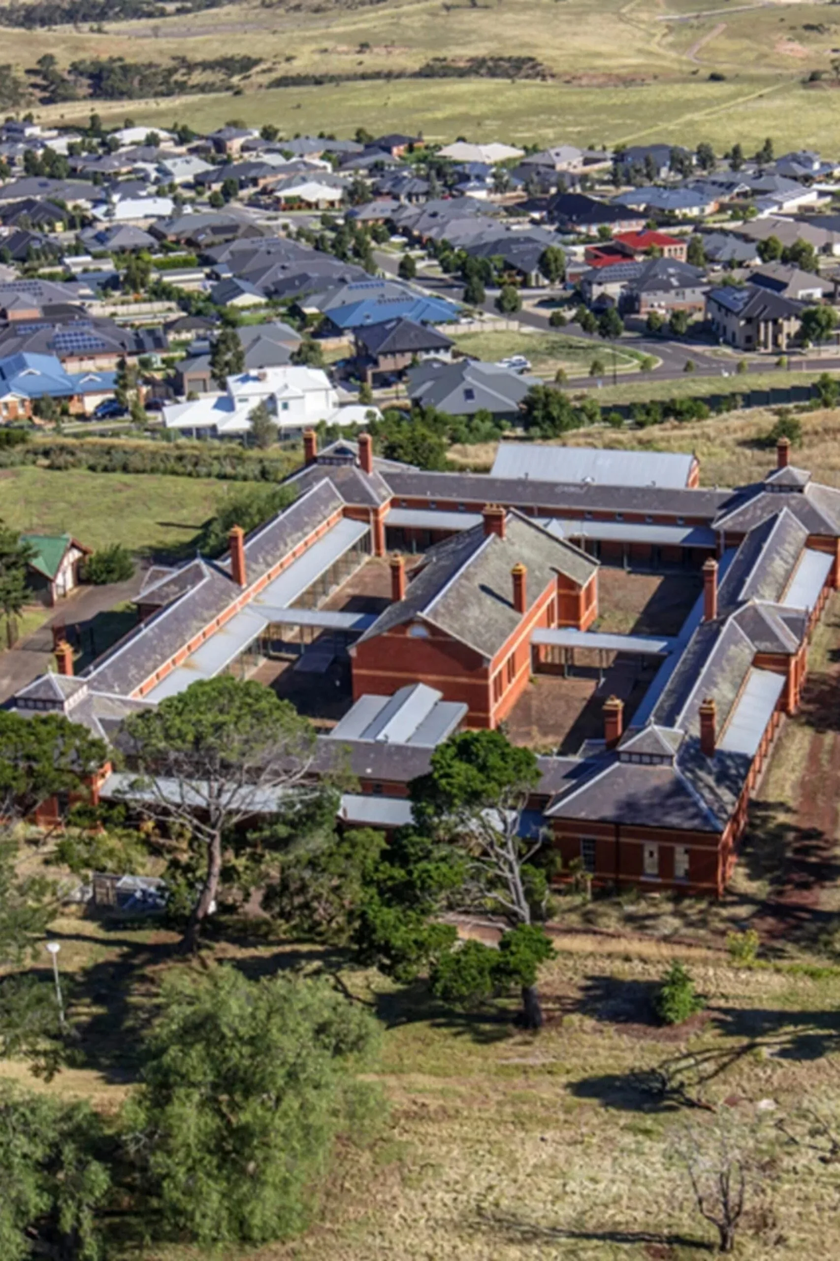 Aerial view of a historic bluestone building complex with orange rooftops, surrounded by trees and suburban houses. The Block 2027 location.