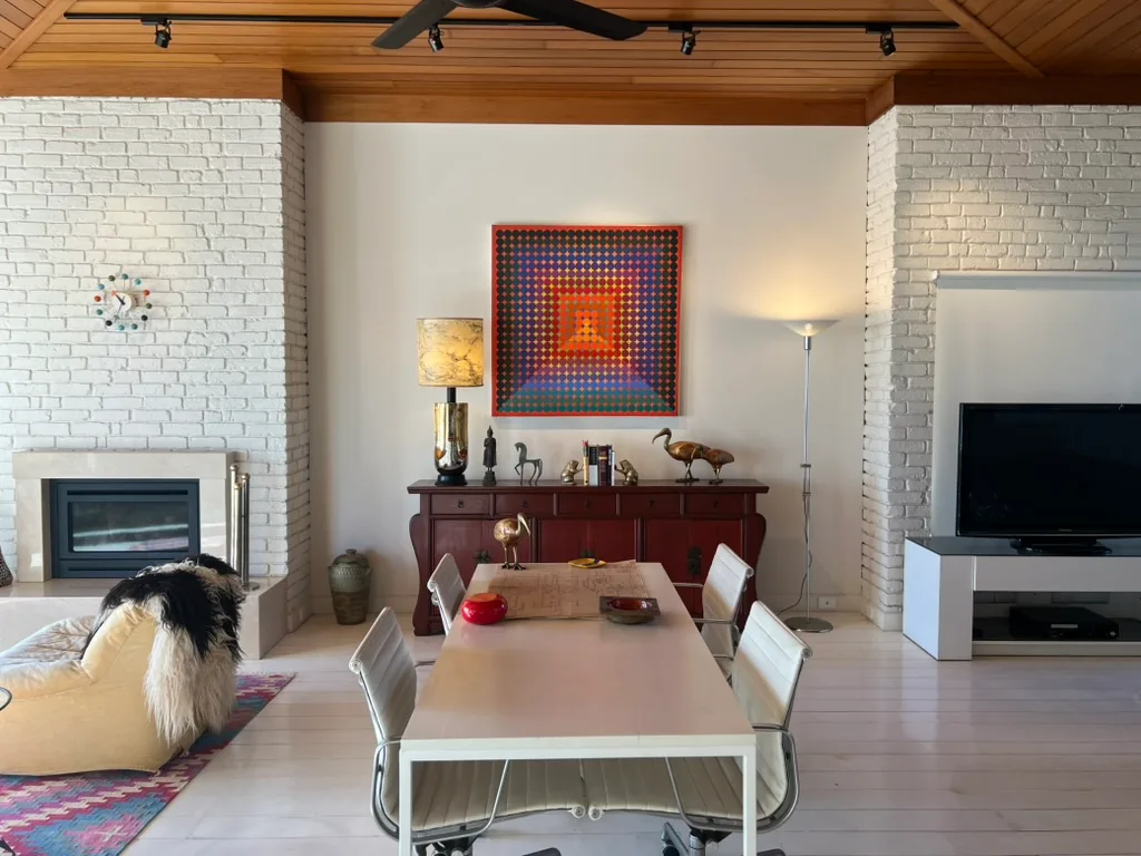 Living room with white brick walls, a dining table, colorful artwork, and a TV on the right Ron Barassi St Kilda home