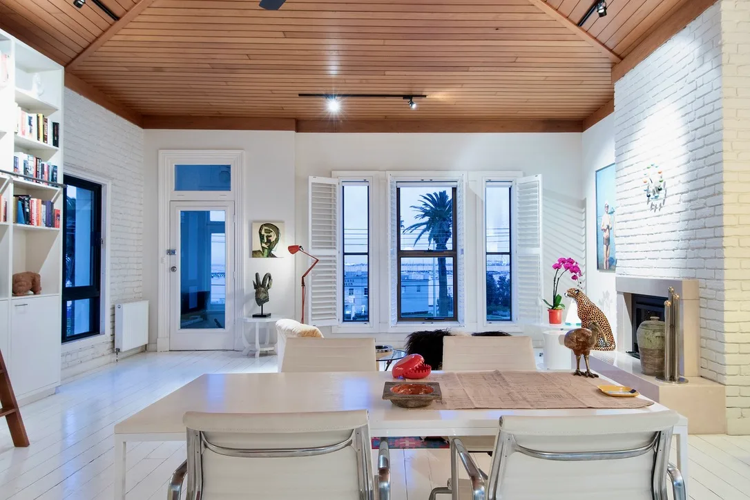 Modern living room with wood ceiling, large windows, white walls, art pieces, and a view of a palm tree outside Ron Barassi St Kilda home
