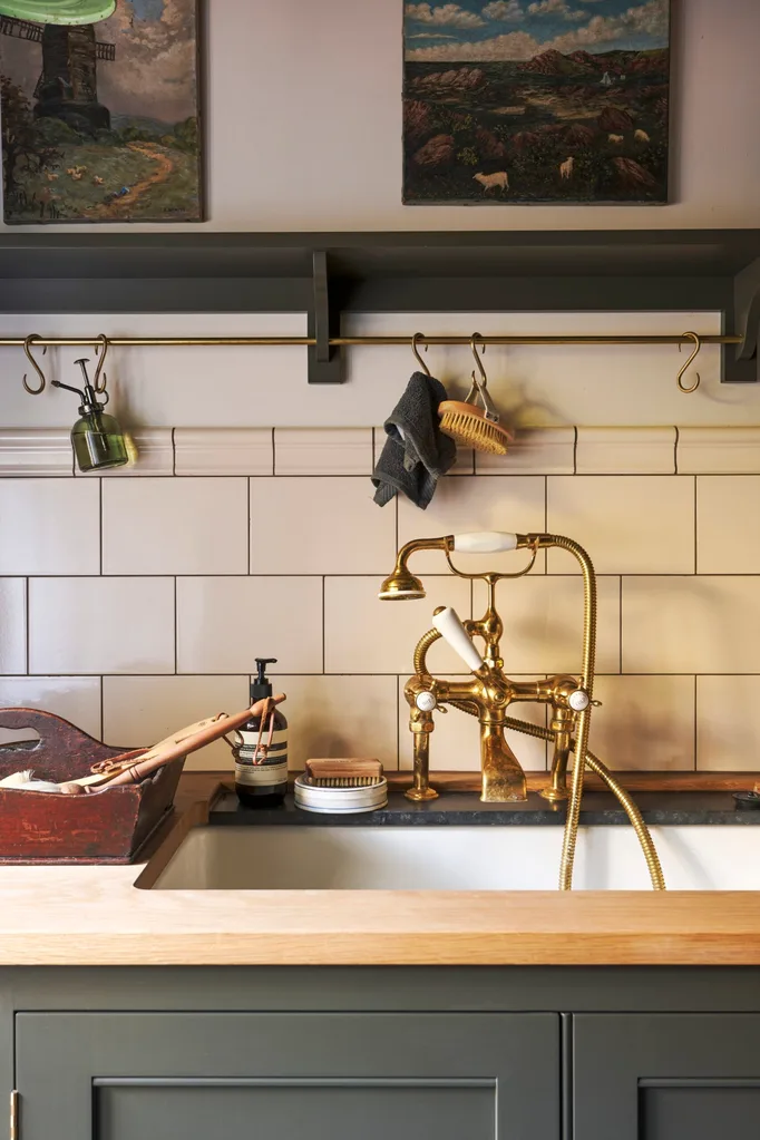 A traditional laundry and mudroom with a timber benchtop, brass tap, tiled splashback, metal hanging rail and a charcoal display shelf above a basin that matches the cupboards below.
