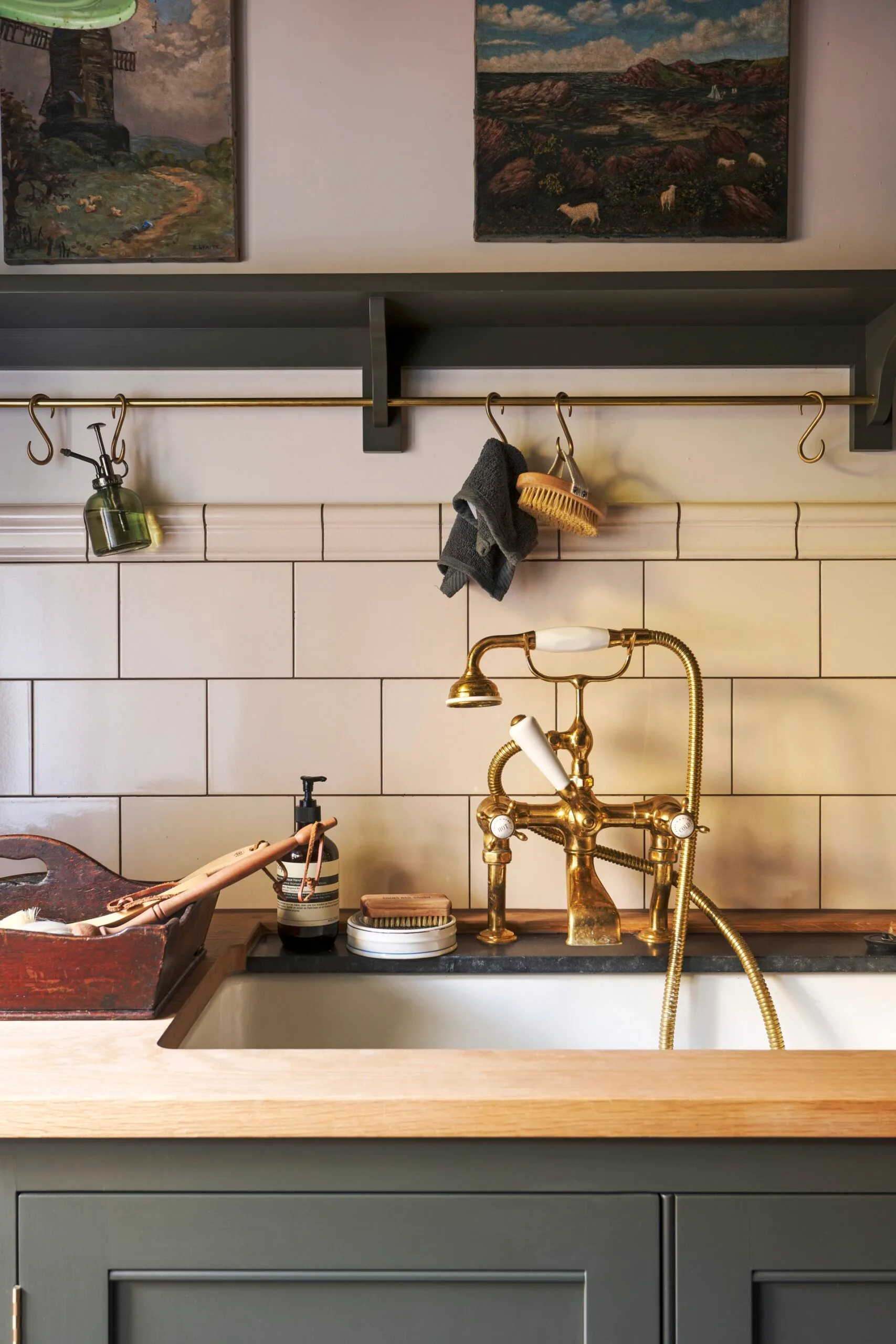 A traditional laundry and mudroom with a timber benchtop, brass tap, tiled splashback, metal hanging rail and a charcoal display shelf above a basin that matches the cupboards below.